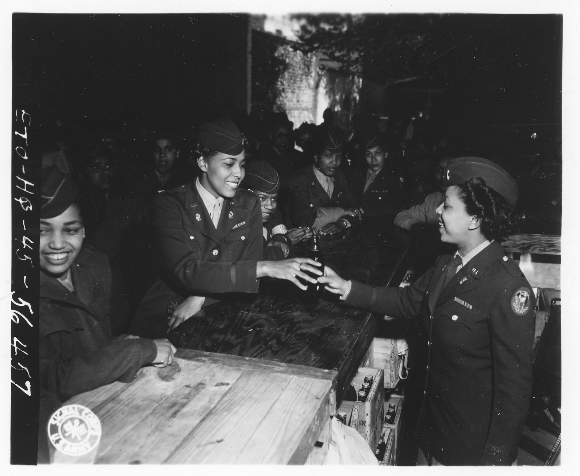 Une femme officier tend une bouteille de Coca-Cola par-dessus un comptoir de bois à une autre, entourées de soldates en uniforme ; des caisses de bouteilles sont empilées au sol