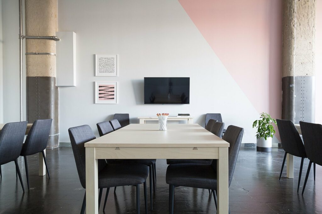 Une salle de réunion vide : une longue table claire, des chaises sombres repoussées, un écran éteint au mur.