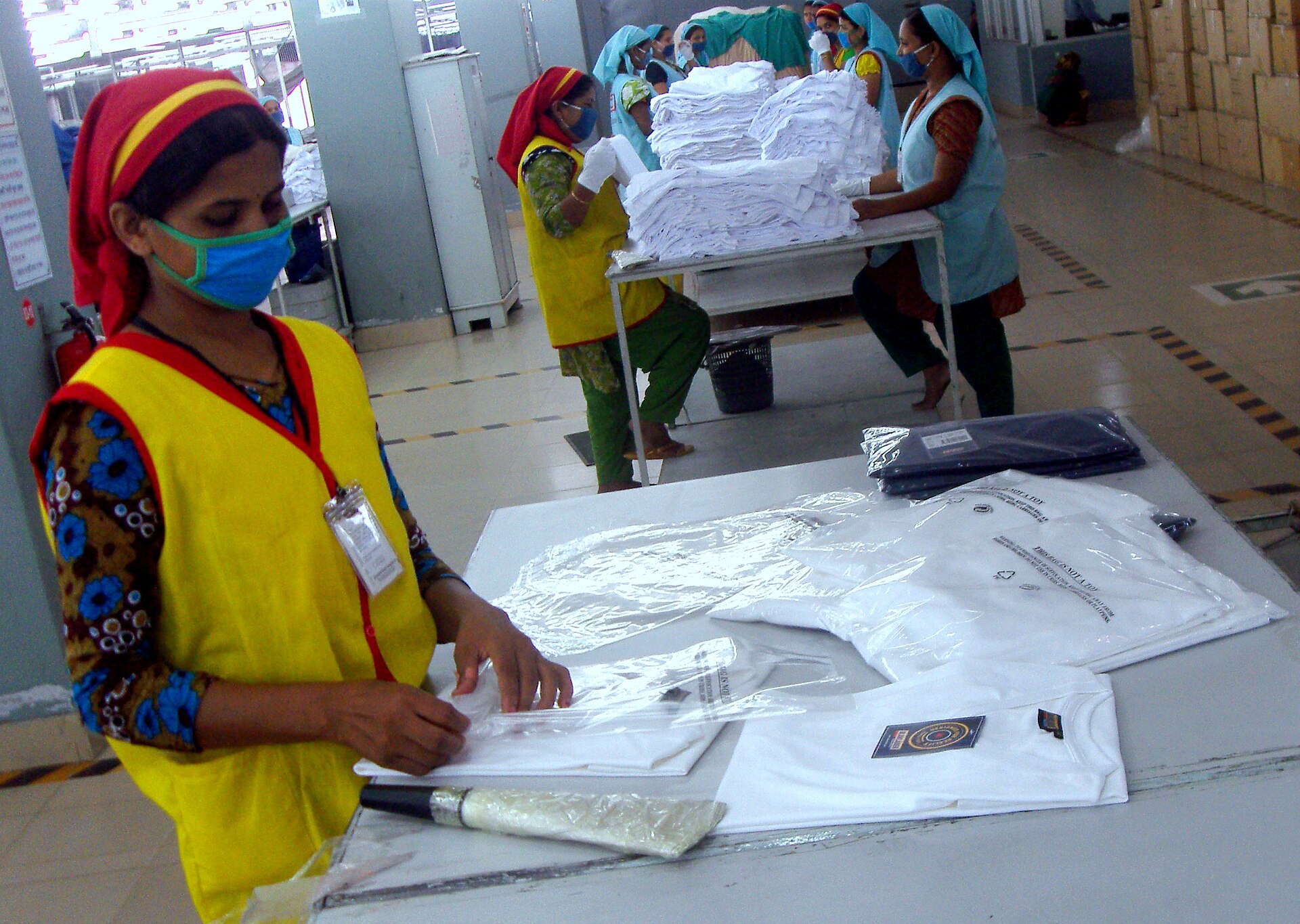 Une ouvriere en gilet jaune controle des tee-shirts blancs sur une table dans une usine de confection.