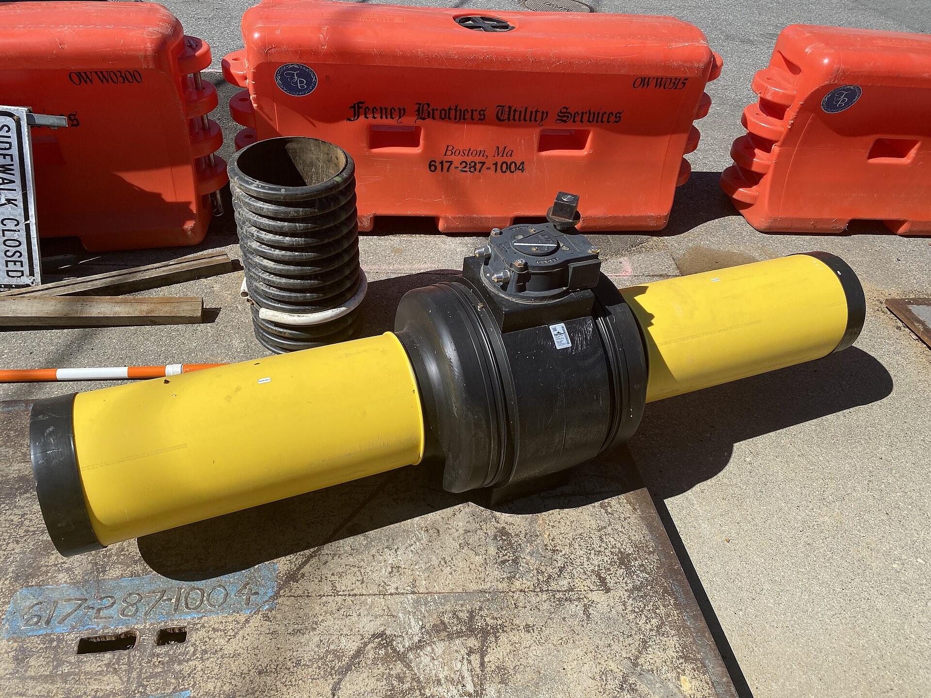 A large black valve mounted on a yellow pipe, resting on a sidewalk before installation.