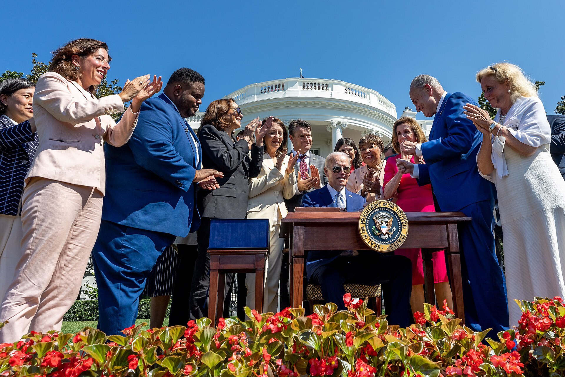 Joe Biden signs a bill at a table on the White House lawn, surrounded by applauding lawmakers.