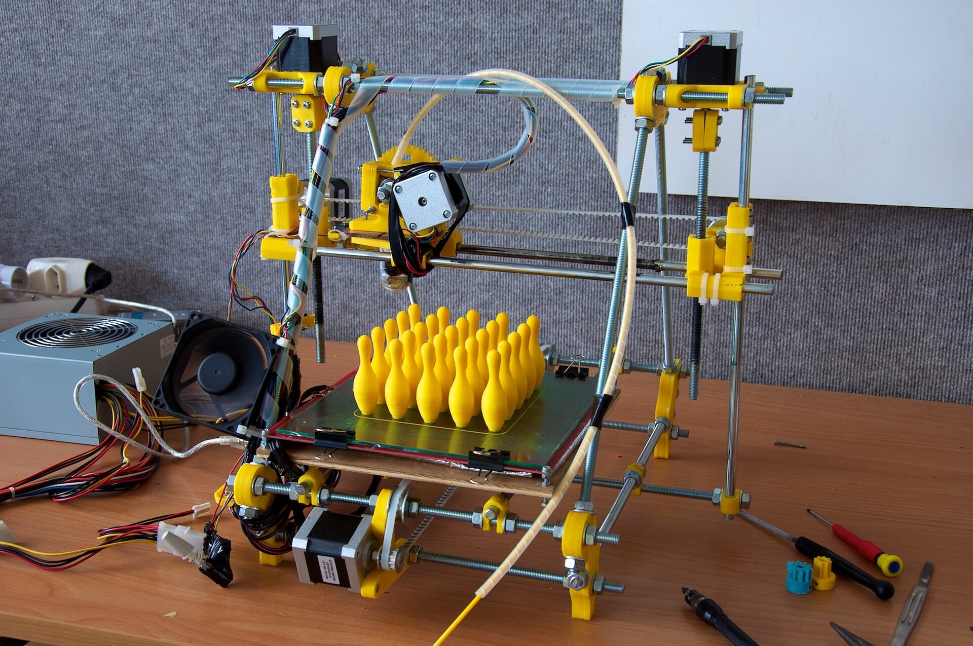 Imprimante 3D en tiges filetées et pièces plastique jaunes, posée sur un établi entre des tournevis, avec une série de petites quilles jaunes imprimées sur son plateau.