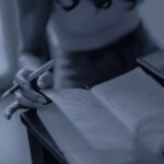 Une main tient un stylo au-dessus d’un carnet ouvert couvert de notes, près d’un ordinateur portable, en bichromie bleu sombre