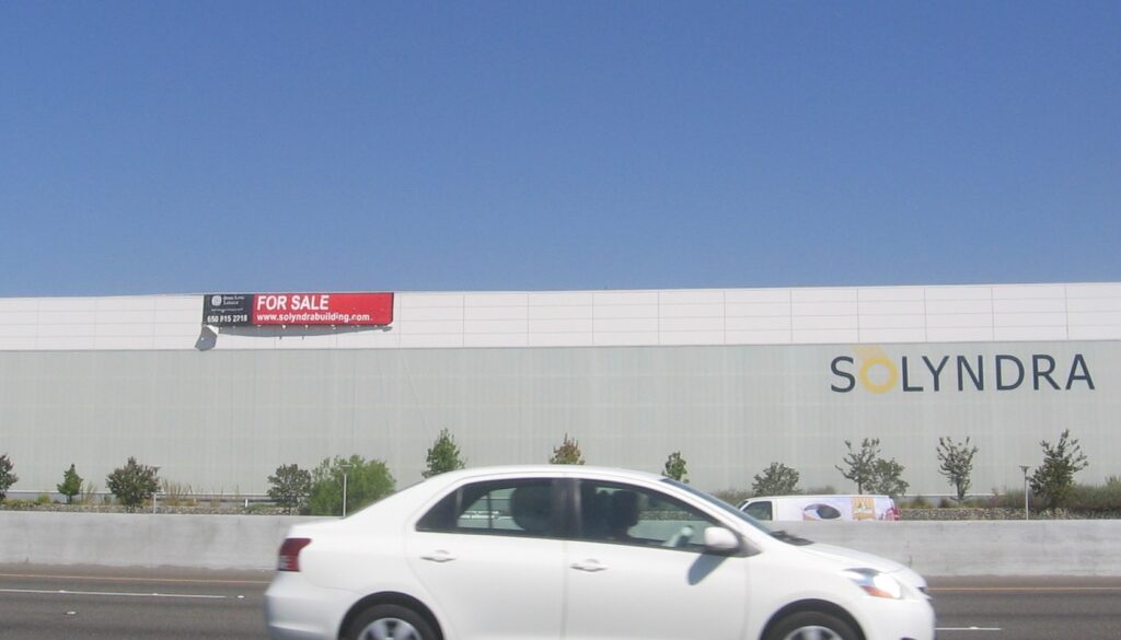 Le bâtiment de Solyndra à Fremont, en Californie, vu depuis l'autoroute, avec un panneau « for sale » posé sur la façade et l'enseigne de l'entreprise encore en place