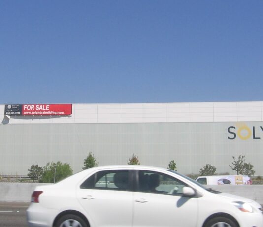 Quand l’État se retire, le risque passe sur votre bilan Le bâtiment de Solyndra à Fremont, en Californie, vu depuis l'autoroute, avec un panneau « for sale » posé sur la façade et l'enseigne de l'entreprise encore en place